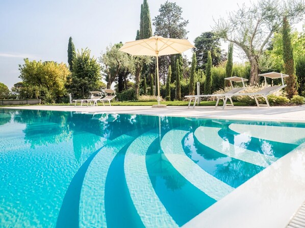 Une piscine d’un bleu éclatant, avec des bords aux motifs ondulés, entourée de chaises longues et de parasols, et située dans un jardin aménagé, agrémenté d’arbres et d’arbustes.