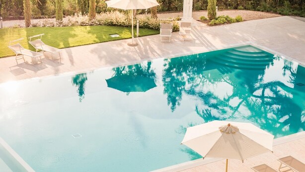 Una piscina rectangular con agua azul cristalina, rodeada por una terraza de madera. Hay sillas de descanso y sombrillas en la terraza.