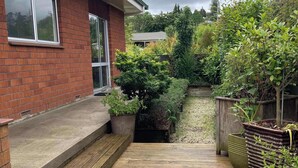 En träaltan som leder fram till ett hus med tegelväggar och ett fönster, omgivet av olika växter och träd.