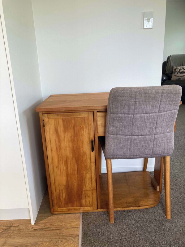 A wooden desk with a grey upholstered chair, a small cabinet, and a light switch on the wall.
