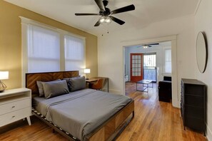 Une chambre à coucher comprenant un lit, des tables de chevet, un bureau, une chaise, un ventilateur de plafond et une porte donnant sur une autre pièce.