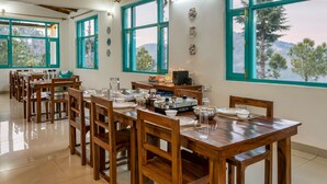 A dining area with wooden tables and chairs, large windows with teal frames, and a view of trees and hills. - TRH Ekuketh (Bhikyasain)