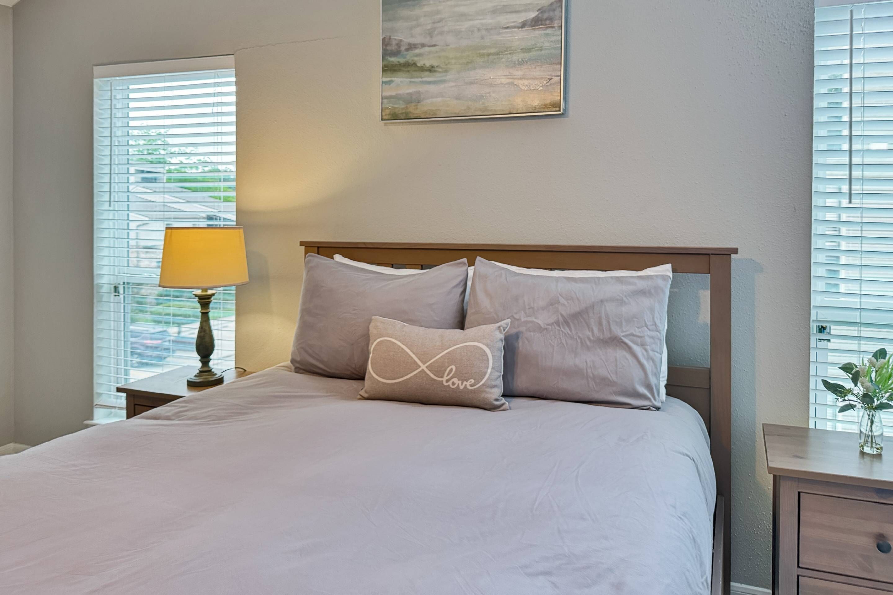 A neatly made bed with a white comforter and pillows, a bedside lamp, a small table with a vase, and a painting on the wall.