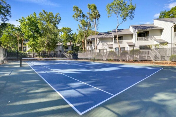 Un court de tennis avec un filet, entouré d’arbres et d’une clôture, avec des bâtiments en arrière-plan.