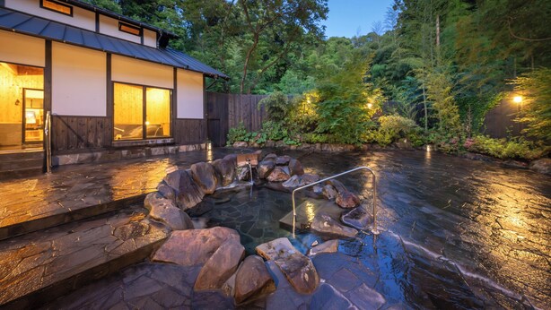 A spa area with a hot tub surrounded by rocks, a wooden deck, and a building with large windows. - Tsuwabuki-no-Hana (Higashisonogi)
