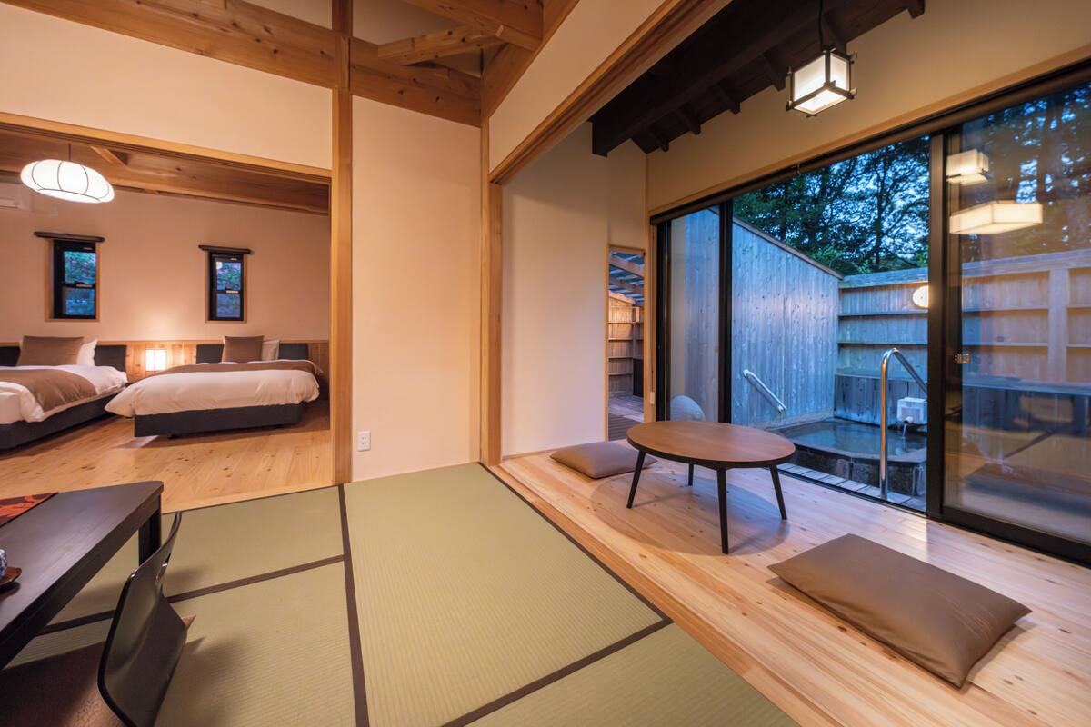 A modern room with a wooden floor, tatami mats, a bed, a table, and a view of a wooden deck and trees.