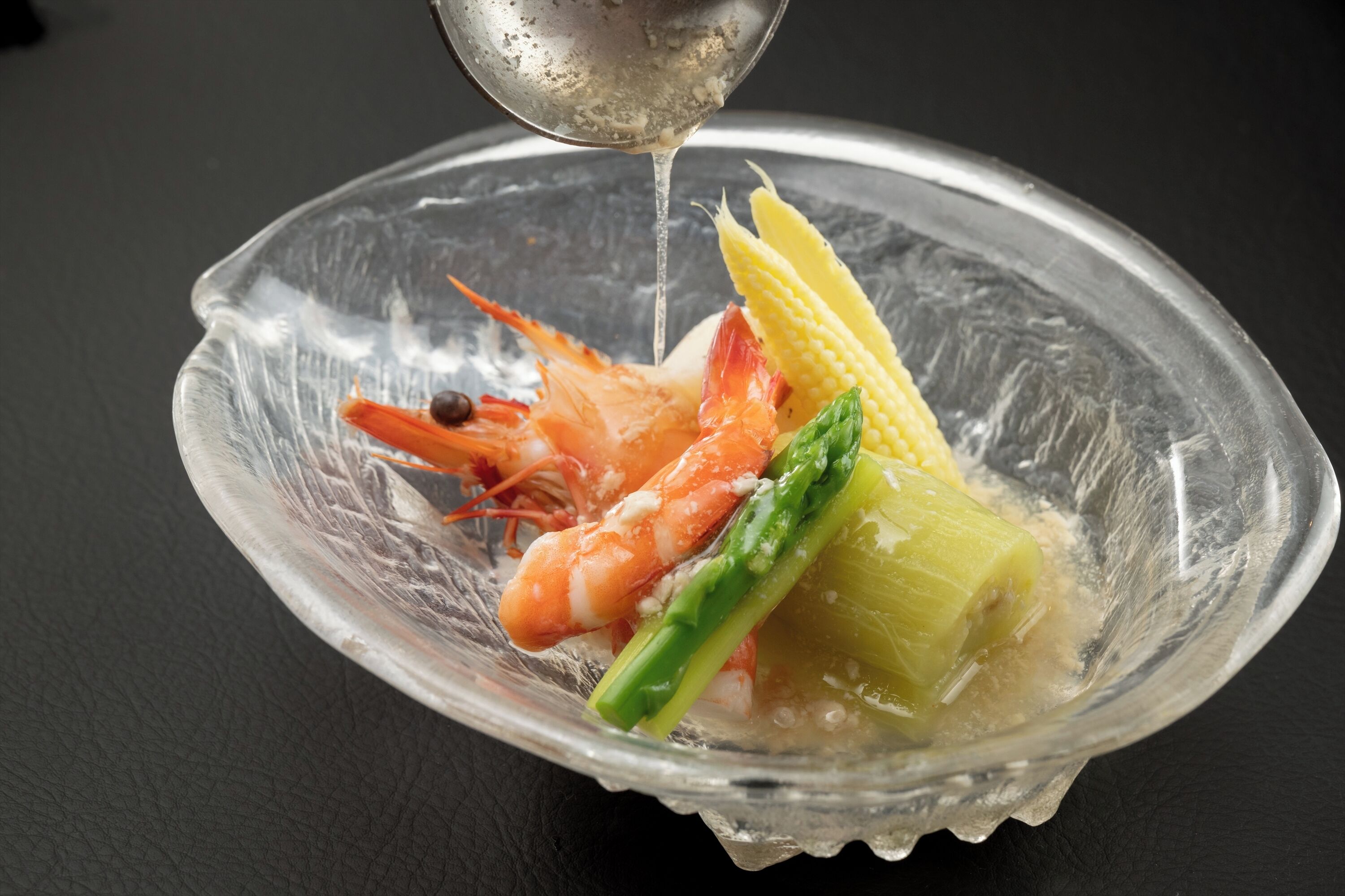 A clear glass bowl containing shrimp, corn, and asparagus on a dark surface.
