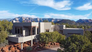 A modern house with a flat roof, large windows, and a balcony. It is surrounded by trees and has a view of mountains in the background.