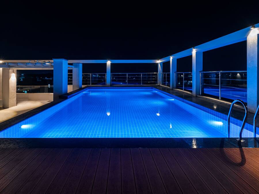 A rooftop swimming pool with a blue-lit interior, surrounded by a wooden deck and metal railings.