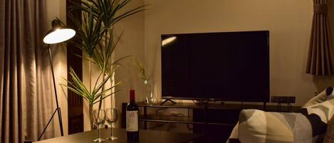 A living room with a flat-screen TV, a table with a bottle and glasses, a lamp, and a sofa with patterned cushions.