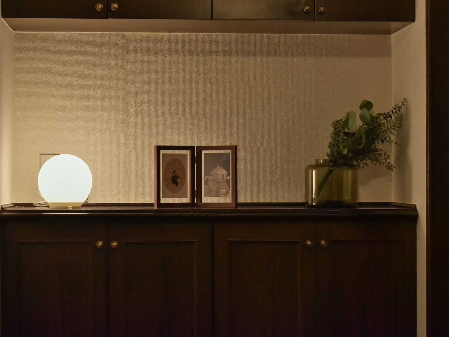 Una habitación con poca luz, un aparador de madera, una lámpara de pared, dos cuadros enmarcados y un florero con una planta.