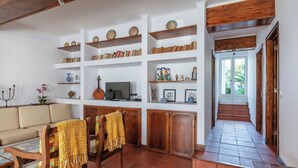 A living room with a sofa, wooden chairs, a TV, shelves with books and decorative items, and a staircase leading to an outdoor area.