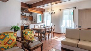 A living room with a sofa, wooden chairs, a dining table, and a kitchen in the background.