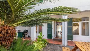 A patio with a wooden table and a grill, surrounded by lush greenery and a white building with green shutters.