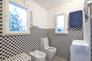 Experience the unique charm of this bathroom, featuring a checkered tile pattern, toilet, bidet, and washing machine, with two windows offering a view of the outdoors.