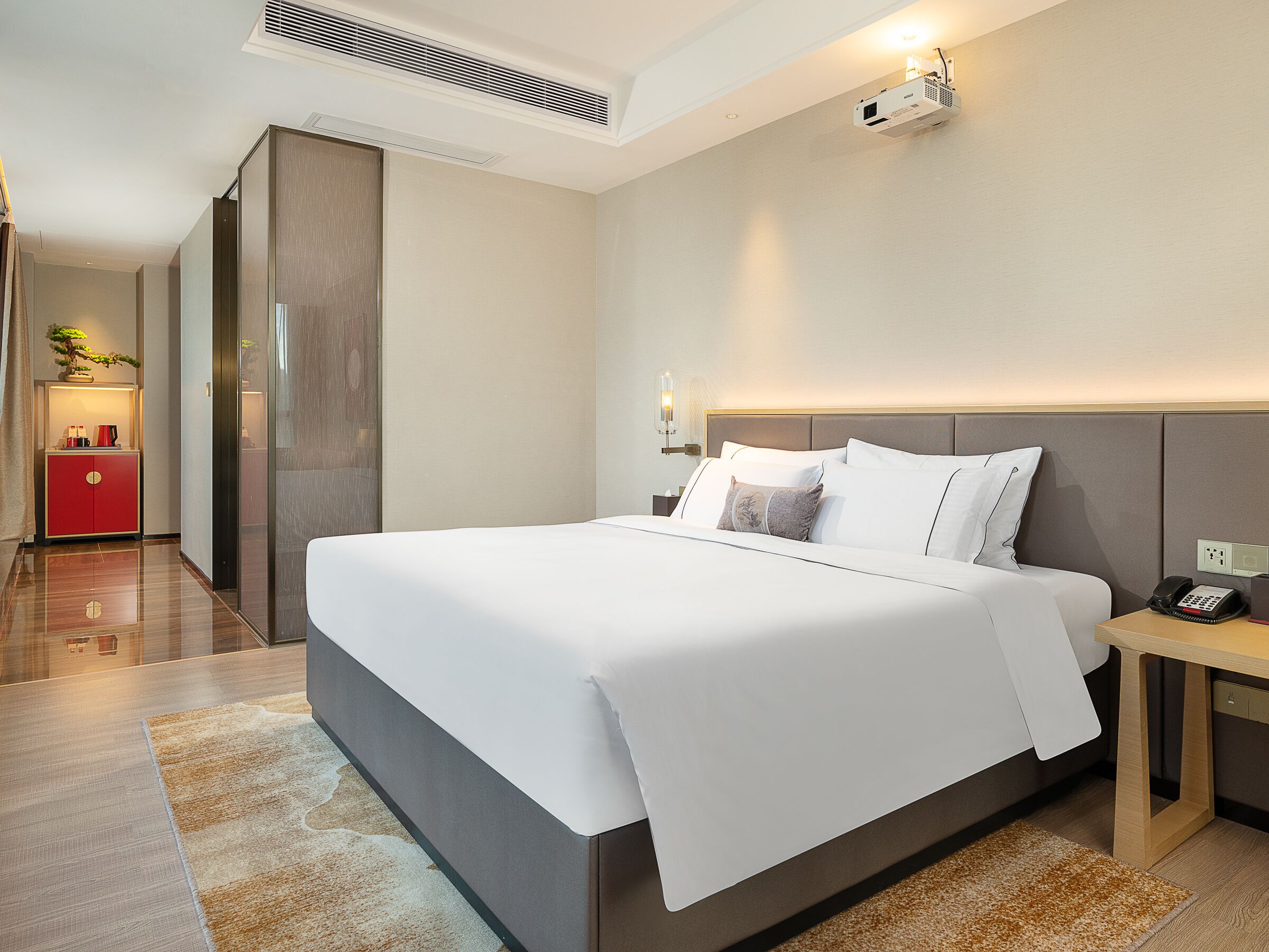 A modern hotel room with a large bed, a bedside table with a phone, a red cabinet, and a ceiling projector.