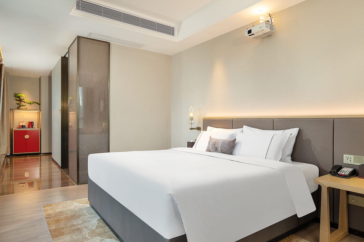 A modern hotel room with a large bed, a bedside table with a phone, a red cabinet, and a ceiling projector.