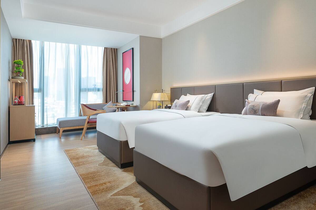 A hotel room with two beds, a desk, a chair, a small table, a lamp, and a large window with curtains.