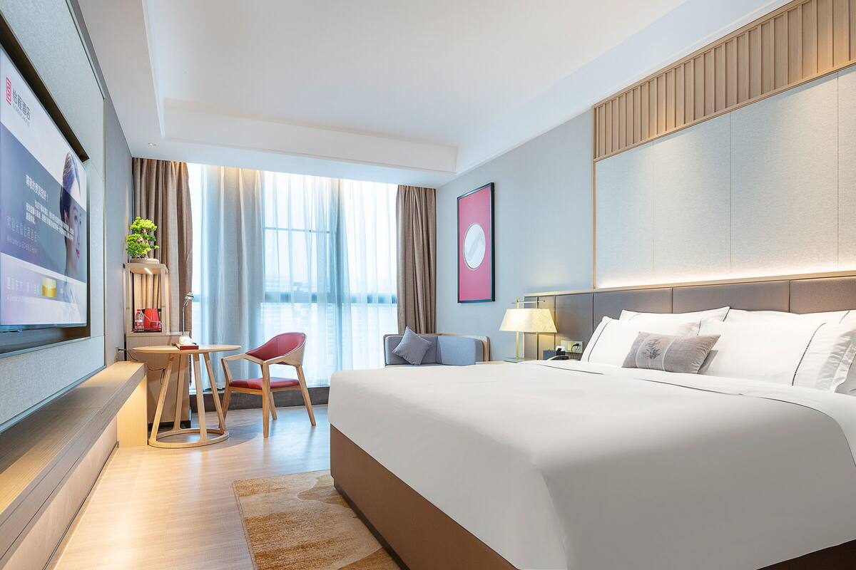 A modern hotel room with a large bed, a desk with a chair, a television, and a window with curtains.