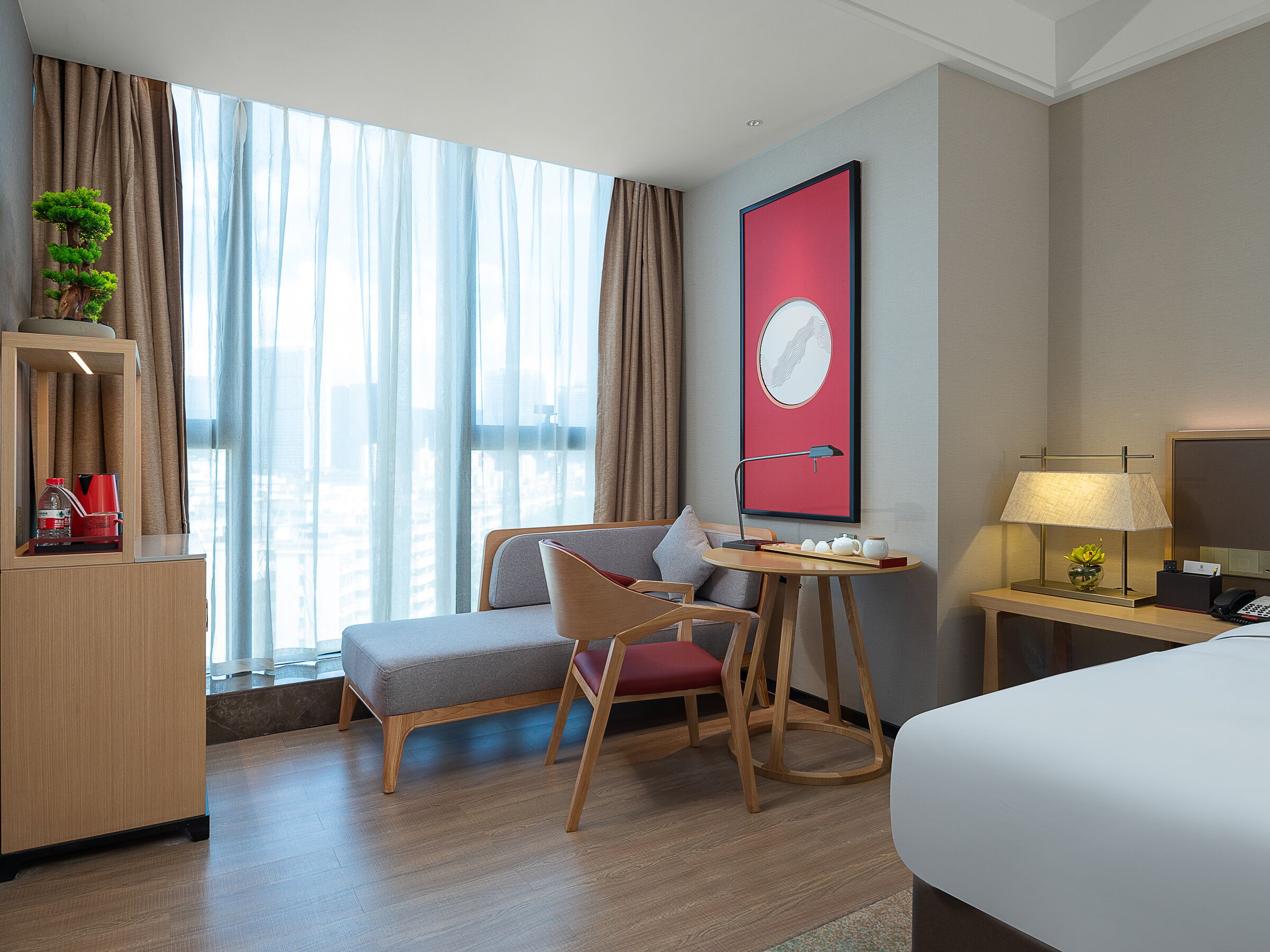 Quarto de hotel com cama, escrivaninha com luminária, cadeira, pequena mesa com xícaras, planta e vista da cidade por grandes janelas.