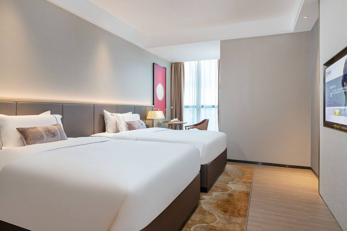 A hotel room with two beds, a nightstand, a lamp, a chair, and a large window with curtains.
