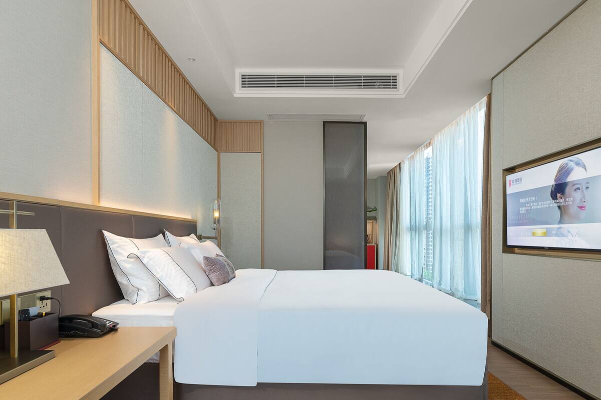 A modern hotel room with a large bed, a desk, a TV, and a window with curtains.