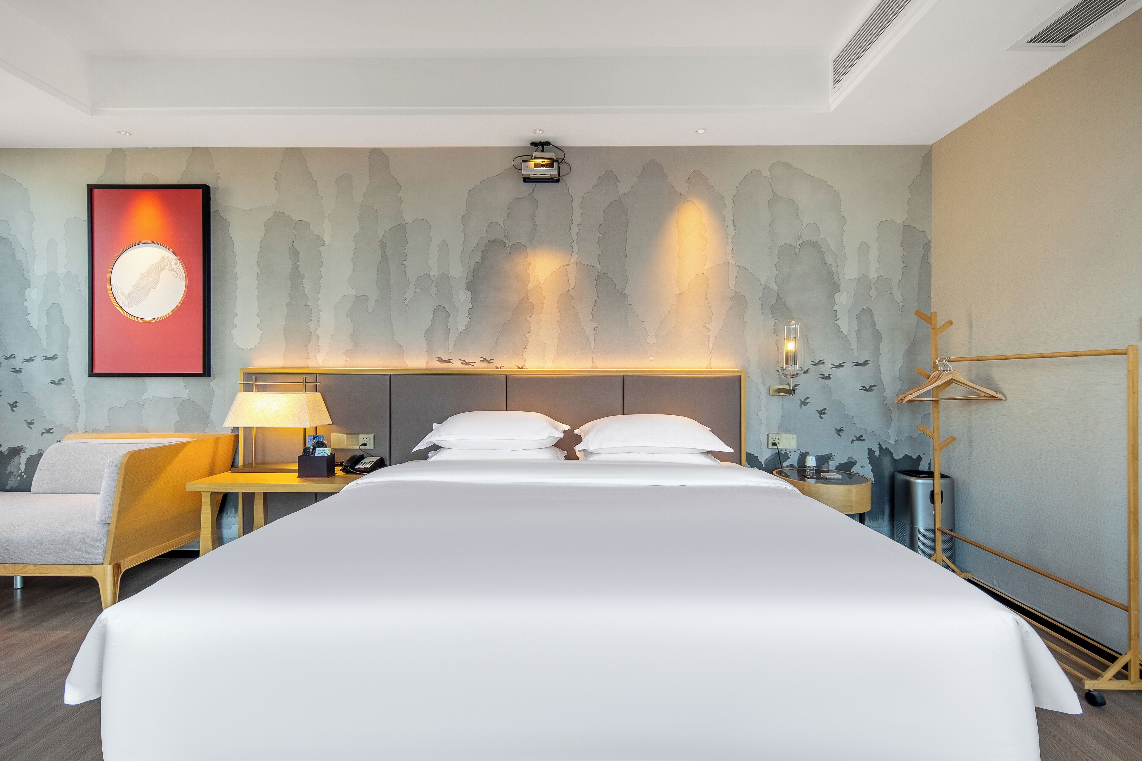 Une chambre d’hôtel avec un grand lit, une table de chevet avec une lampe, un projecteur fixé au mur et un porte-manteau.