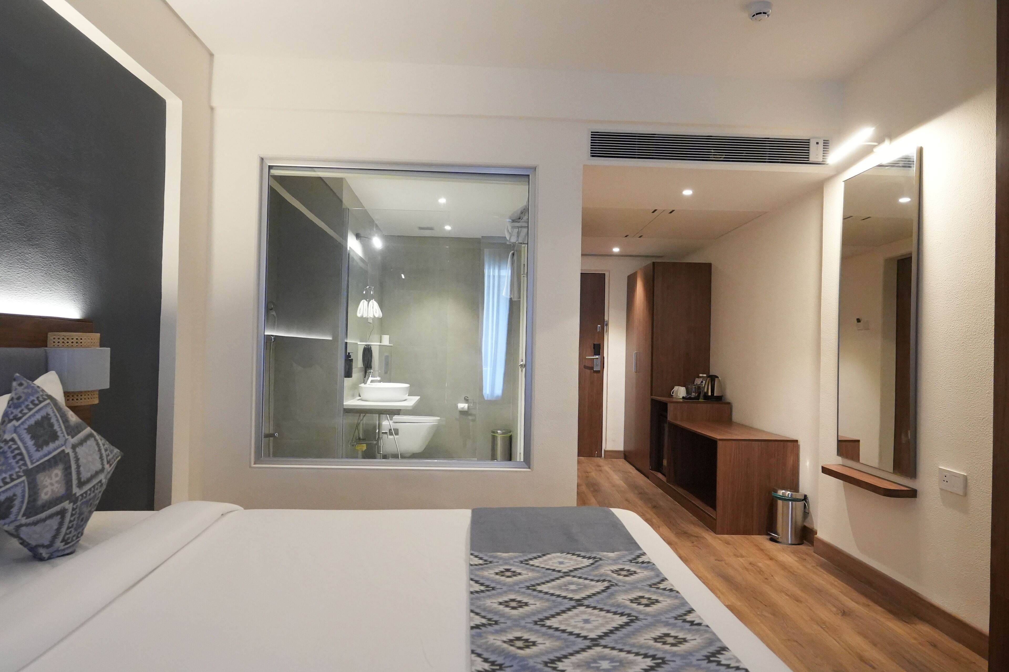 A modern hotel room with a large bed, a bedside table, a window with a view of the bathroom, and a small mirror on the wall.