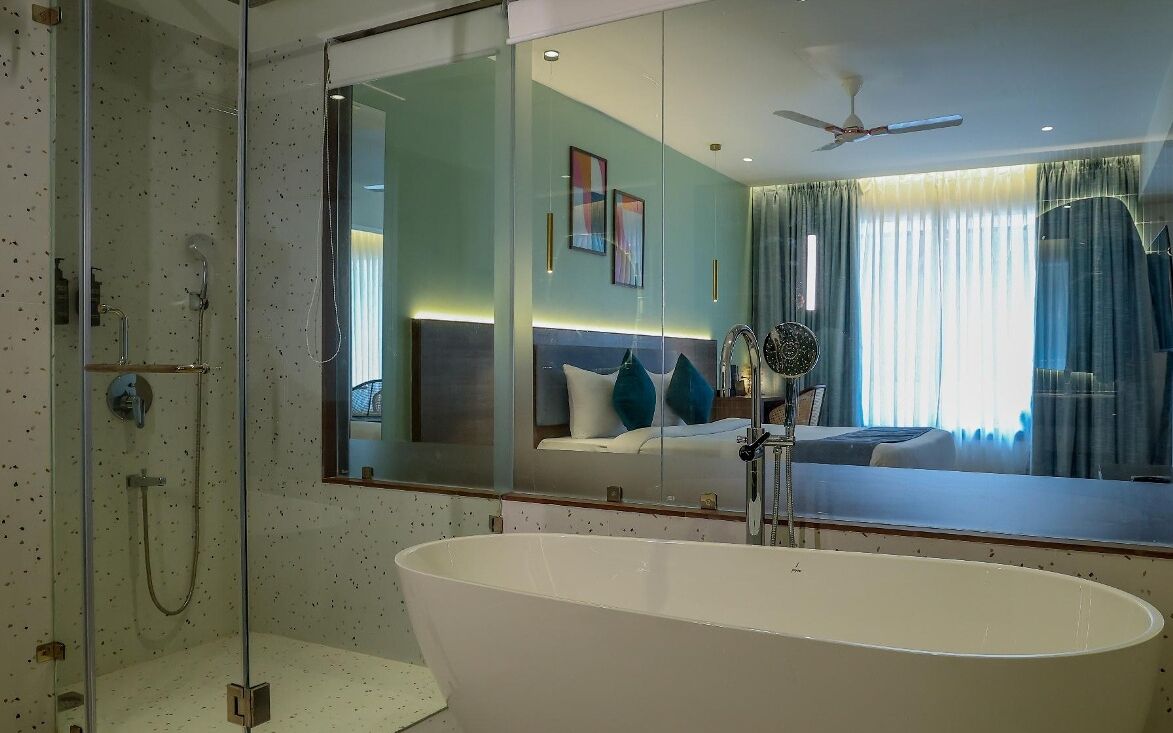 A modern bathroom with a freestanding bathtub, a glass-enclosed shower, and a large mirror reflecting a bedroom with a bed and curtains.