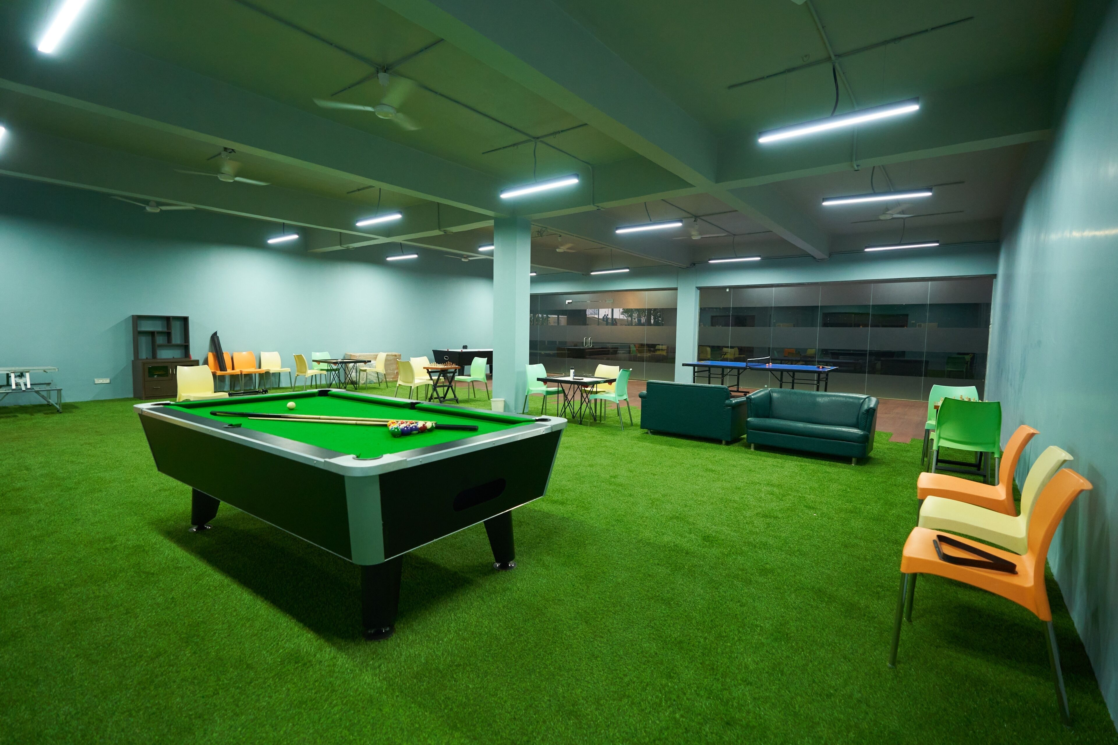 A pool table with a green felt surface, surrounded by artificial grass, and a green sofa in a room with a high ceiling and fluorescent lighting.