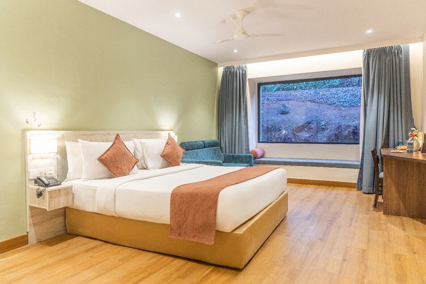 A hotel room with a bed, a desk, a chair, a nightstand, and a window with a view of a rocky landscape.