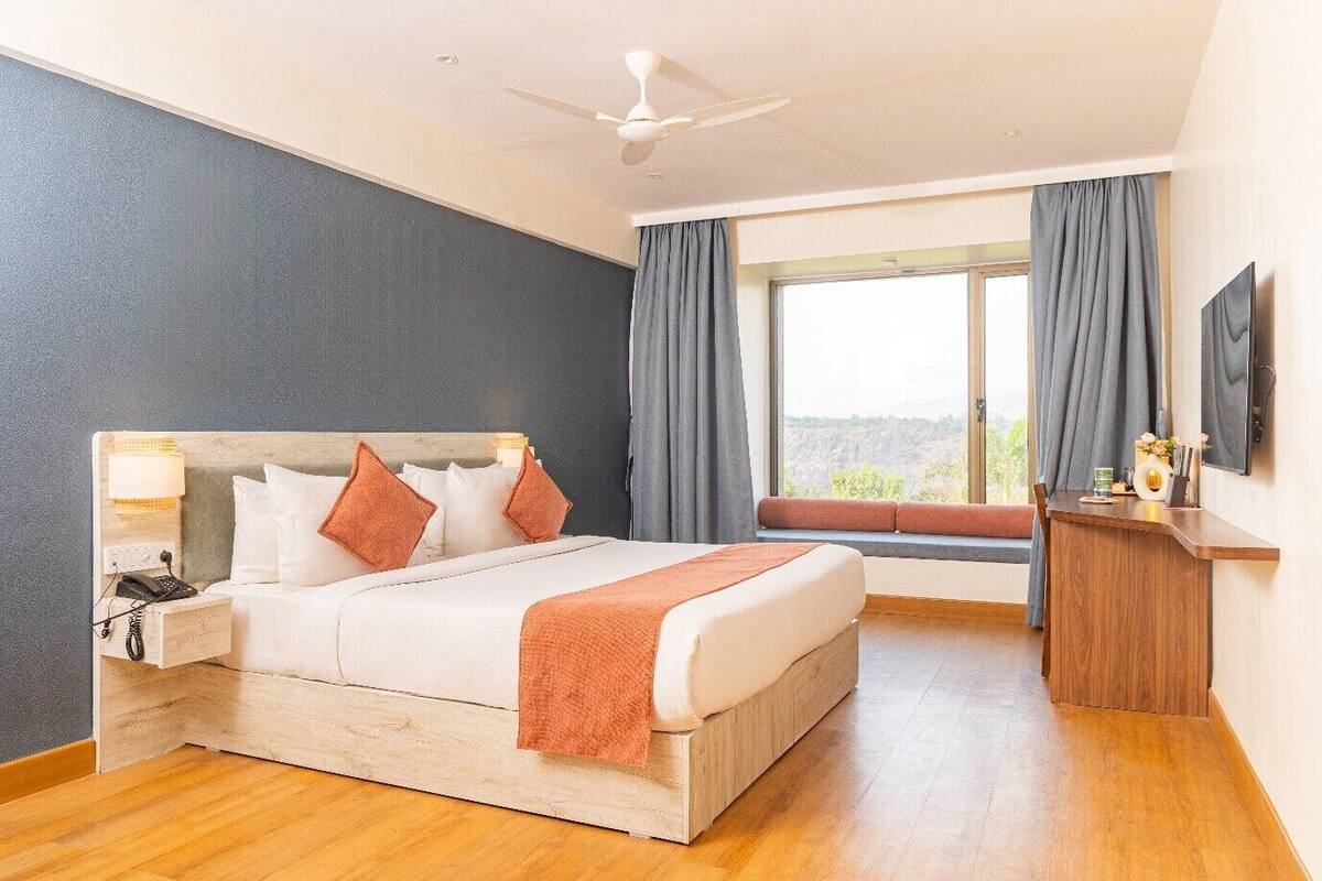 A hotel room with a bed, bedside lamp, phone, window with curtains, and a TV.