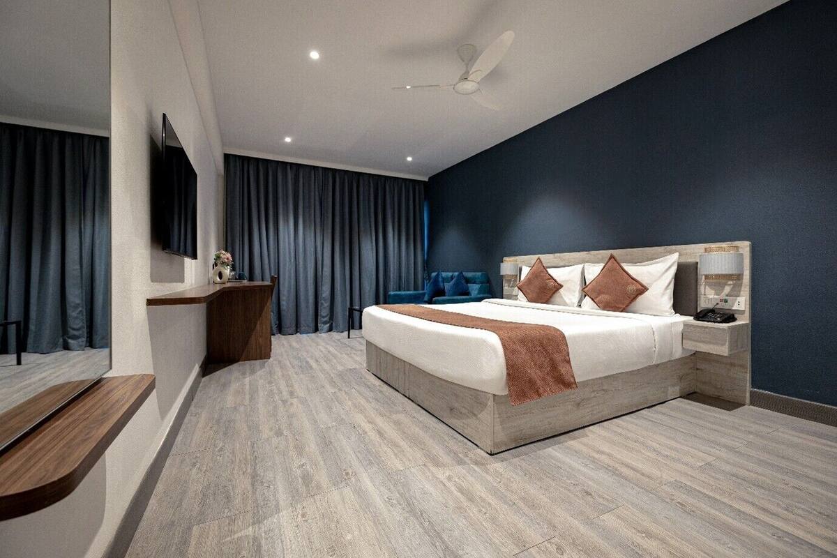 A modern hotel room with a large bed, a desk, a chair, a TV, and a ceiling fan.