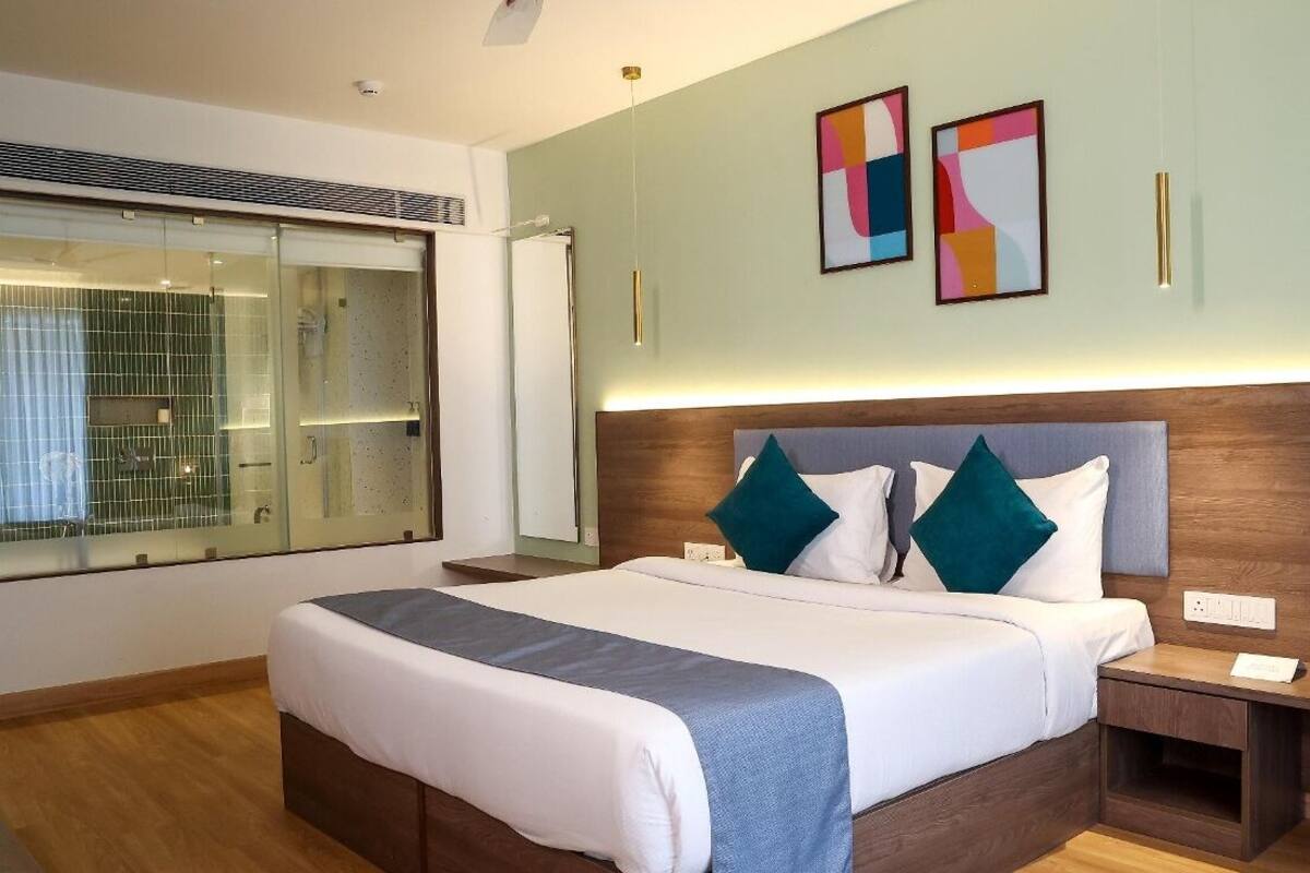 A hotel room with a large bed, a bedside table, a nightstand, a window with a view, and two abstract paintings on the wall.