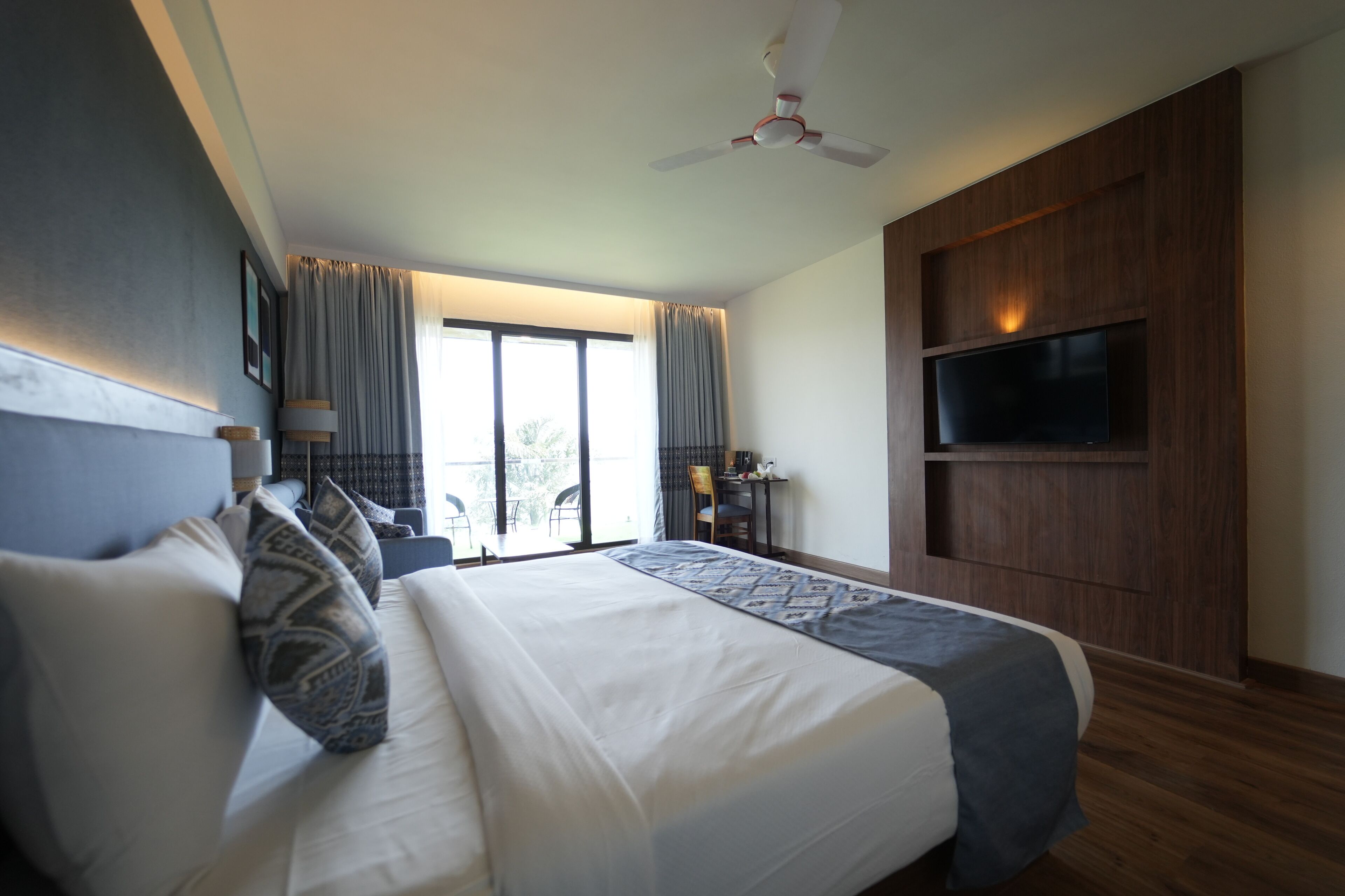 A modern hotel room with a large bed, a ceiling fan, a flat-screen TV mounted on the wall, and a balcony with a view.