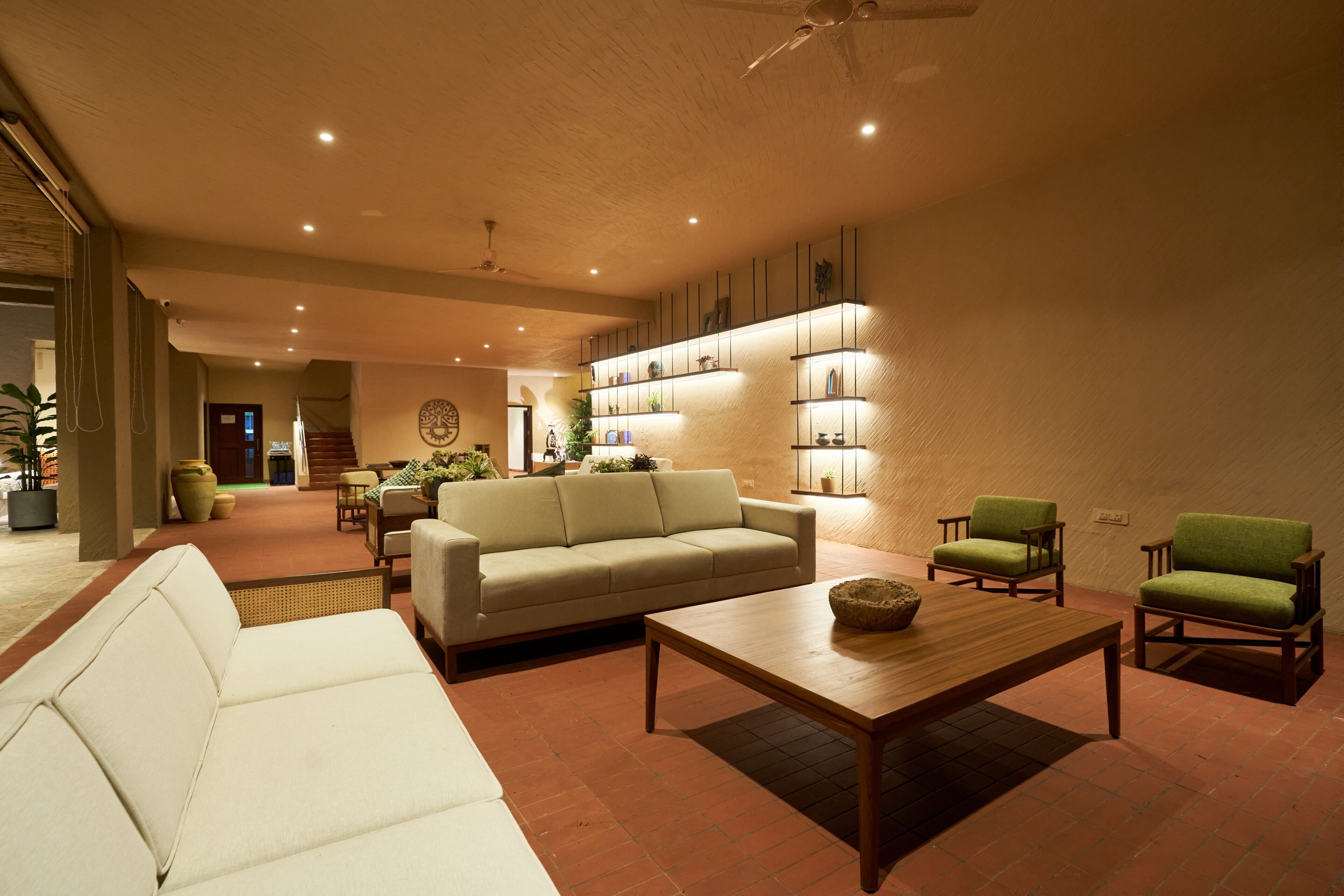 A spacious living area with a sofa, a coffee table, and two armchairs. There is a staircase leading to an upper level, and shelves with decorative items on the wall.