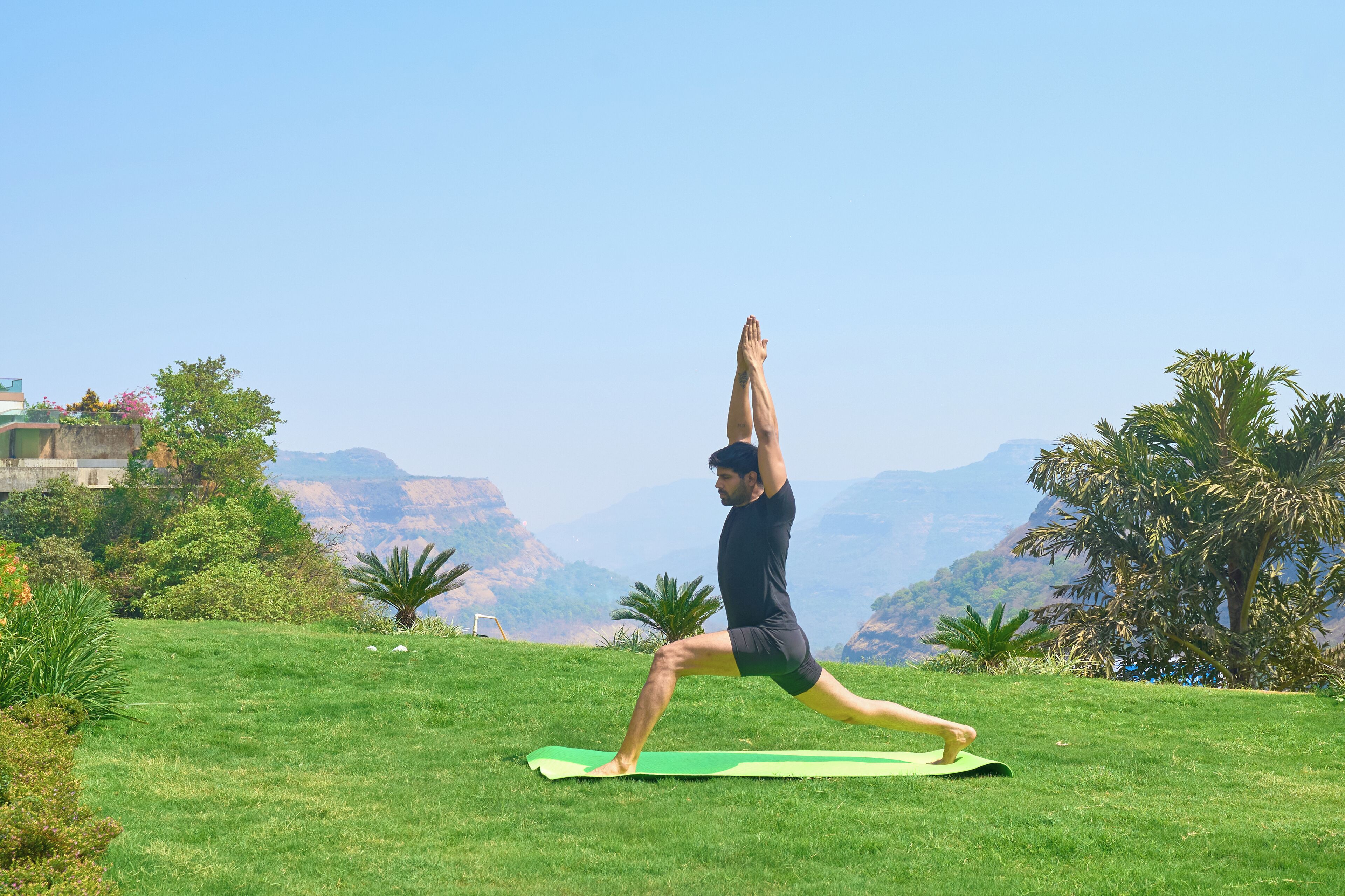 A person performing a yoga pose on a green mat in a grassy outdoor area with a mountainous backdrop.