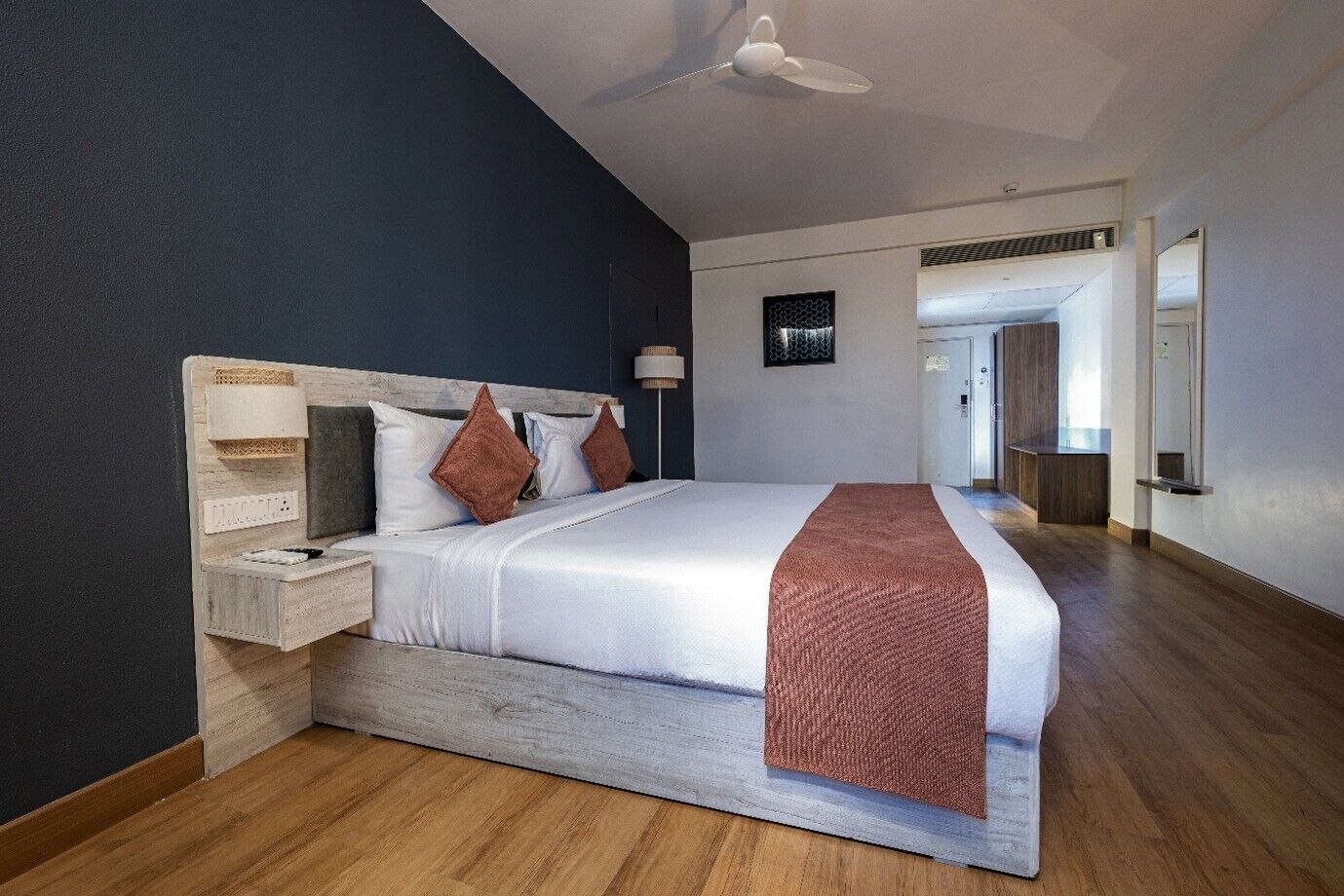 A modern hotel room with a large bed, a wooden headboard, a ceiling fan, and a view of the hallway.