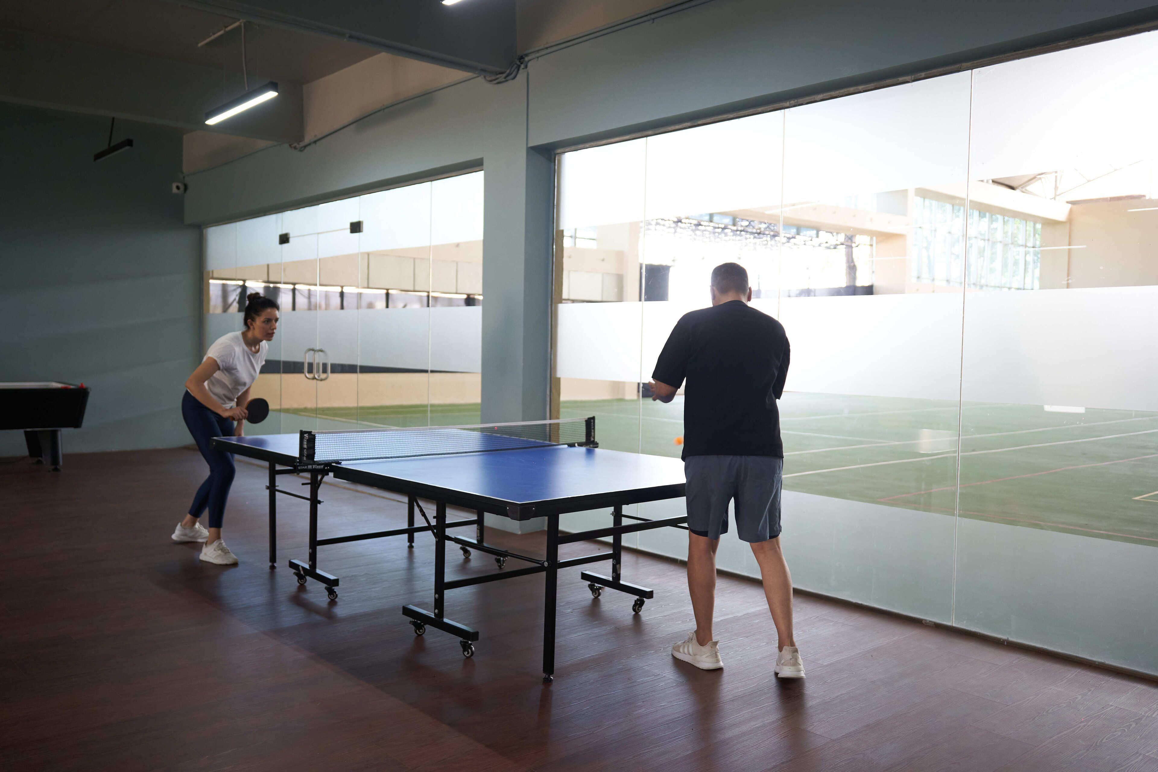 A ping pong table in a room with large windows, a basketball court visible outside, and a piano in the background.
