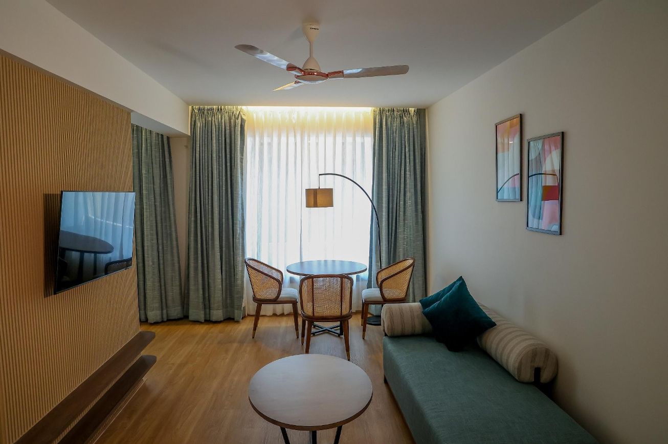 A modern living room with a flat-screen TV mounted on the wall, a ceiling fan, a round dining table with chairs, a sofa, and a window with curtains.