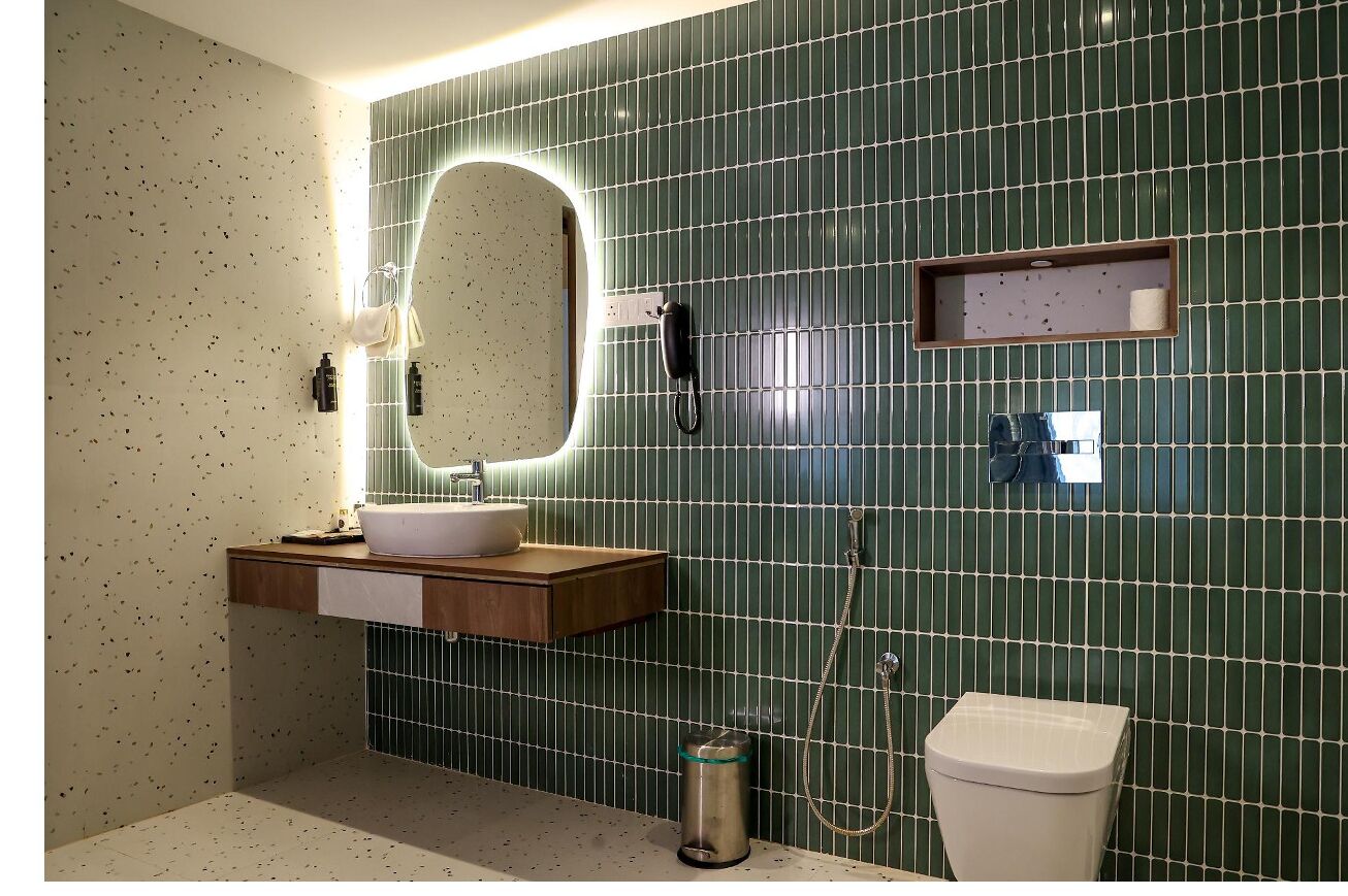 Un baño con pared de azulejos verdes, tocador de madera, lavamanos blanco, espejo, cabezal de ducha manual y retrete.
