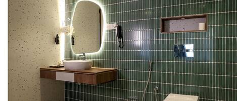 Un baño con pared de azulejos verdes, tocador de madera, lavamanos blanco, espejo, cabezal de ducha manual y retrete.