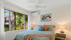 Une chambre avec une grande fenêtre, un lit avec de nombreux oreillers, un ventilateur de plafond et un climatiseur fixé au mur.