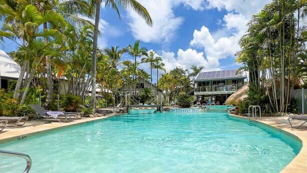 Une piscine aux eaux cristallines, entourée de chaises longues et de palmiers, avec un bâtiment visible au loin.