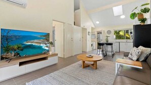 Un salon moderne avec un téléviseur à écran plat, une table basse en bois, un canapé et un coin cuisine à l’arrière-plan.