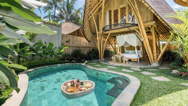 Una piscina con barra flotante, una casa de madera con balcón y exuberante vegetación tropical.