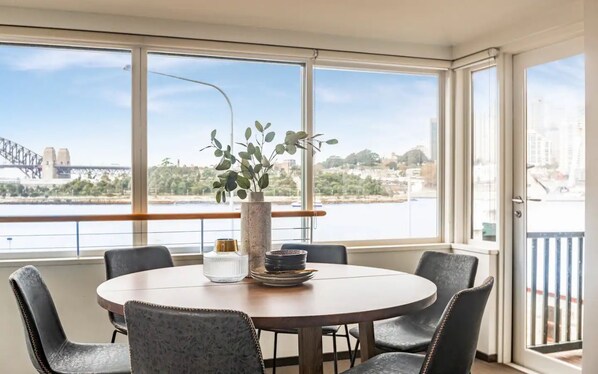 A dining area with a round table, six chairs, a vase with green plants, and a view of a bridge and river. - Balmain Wharf Apartments - #1 (Inner West Council)
