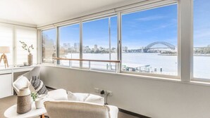 Ein Zimmer mit großem Fenster, das auf einen Fluss und eine Brücke blickt, ein weißes Sofa und ein kleiner Tisch mit Dekorationen.