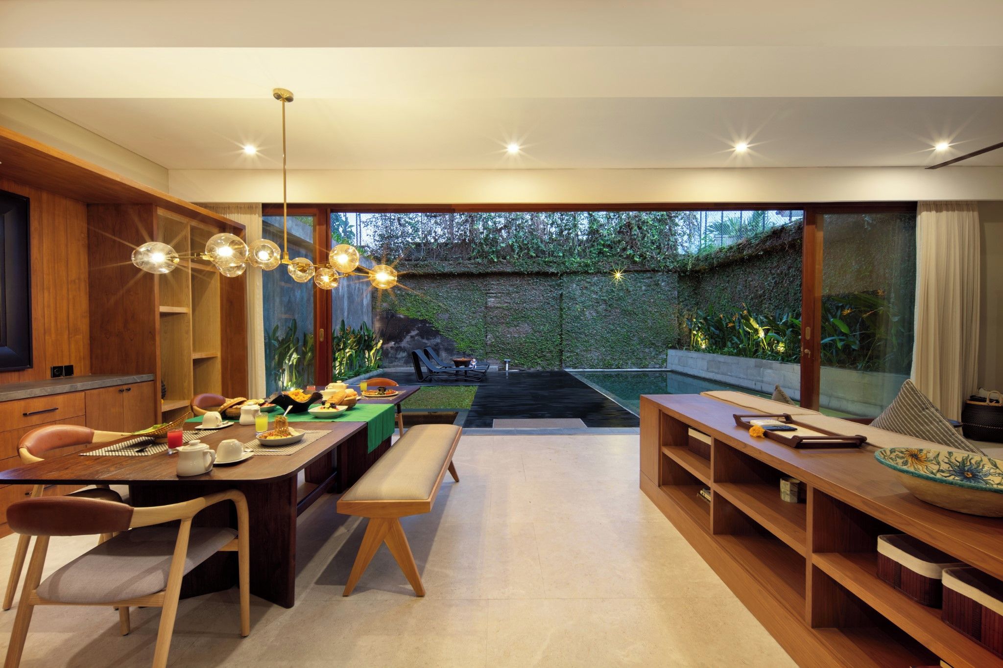 A modern dining area with a wooden table, chairs, and a bench. There is a small pool in the background with a bench and lounge chairs.