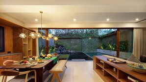 A modern dining area with a wooden table, chairs, and a bench. There is a large window providing a view of a garden and a swimming pool. - Kolila Villa Seminyak by Ini Vie Hospitality (Kerobokan)
